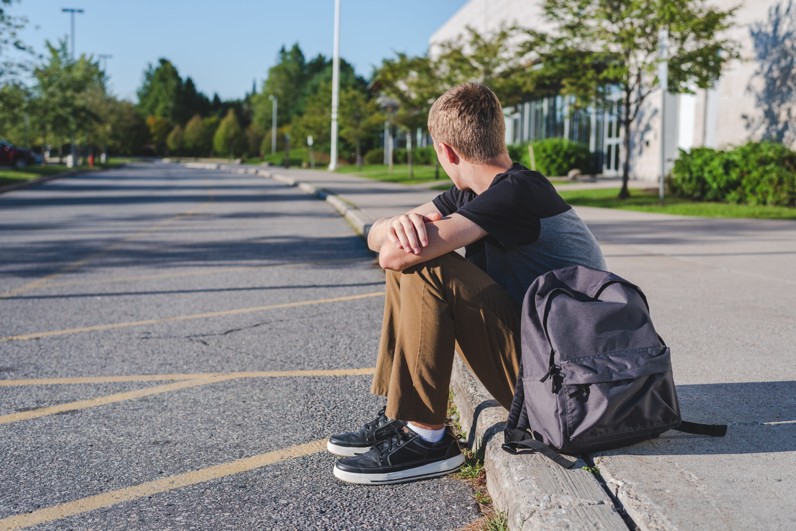 Lonely,Teenage,Boy,Sitting,On,Curb,Next,To,High,School.